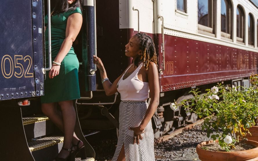 Two women look at each other, and are posed boarding the Napa Valley Wine Train. The woman on the left stands on the stairs to the train's cabin, wearing a green dress. The woman on the right is standing on the ground, weaking a flowy white pattered dress.