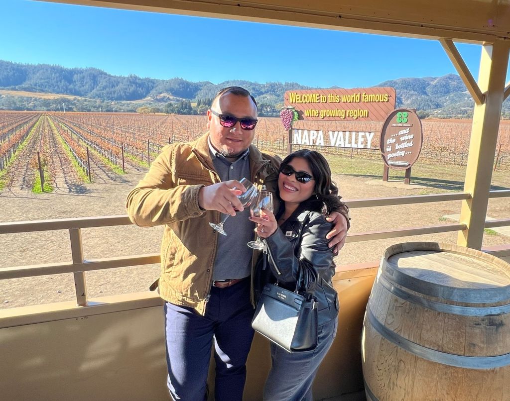 A man and woman wearing sunglasses clink their glasses of red wine together. They are posed in front of the Napa Valley wooden sign and vineyard.