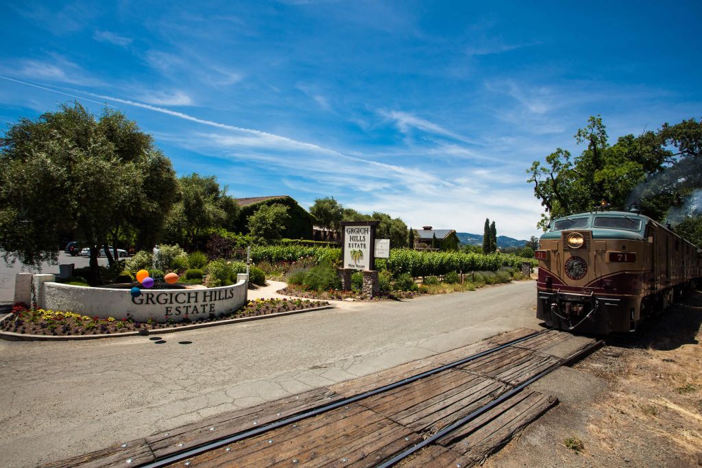 A photo of Grgich Hills Estate and the Napa Valley Wine Train. On the left are two signs that state "Grgich Hills Estate". The leftmost sign has multicolour balloons floating above it. To the right, the Nava Valley Wine Train sits on its track.