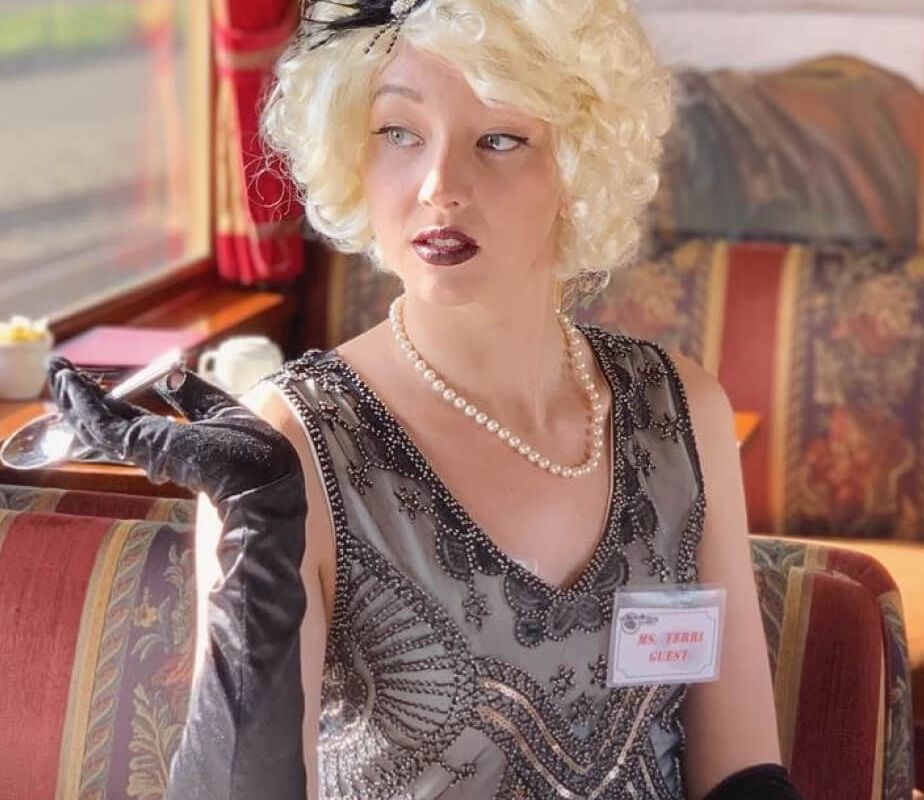 A woman is posed seated in a dining booth of the Napa Valley Wine Train, gazing out the window. She wears a light blonde wig, a grey patterned flapper-style dress, and black opera-length gloves. She holds a metal spoon in her right hand. A name tag is fixed on the chest of her dress, reading 
