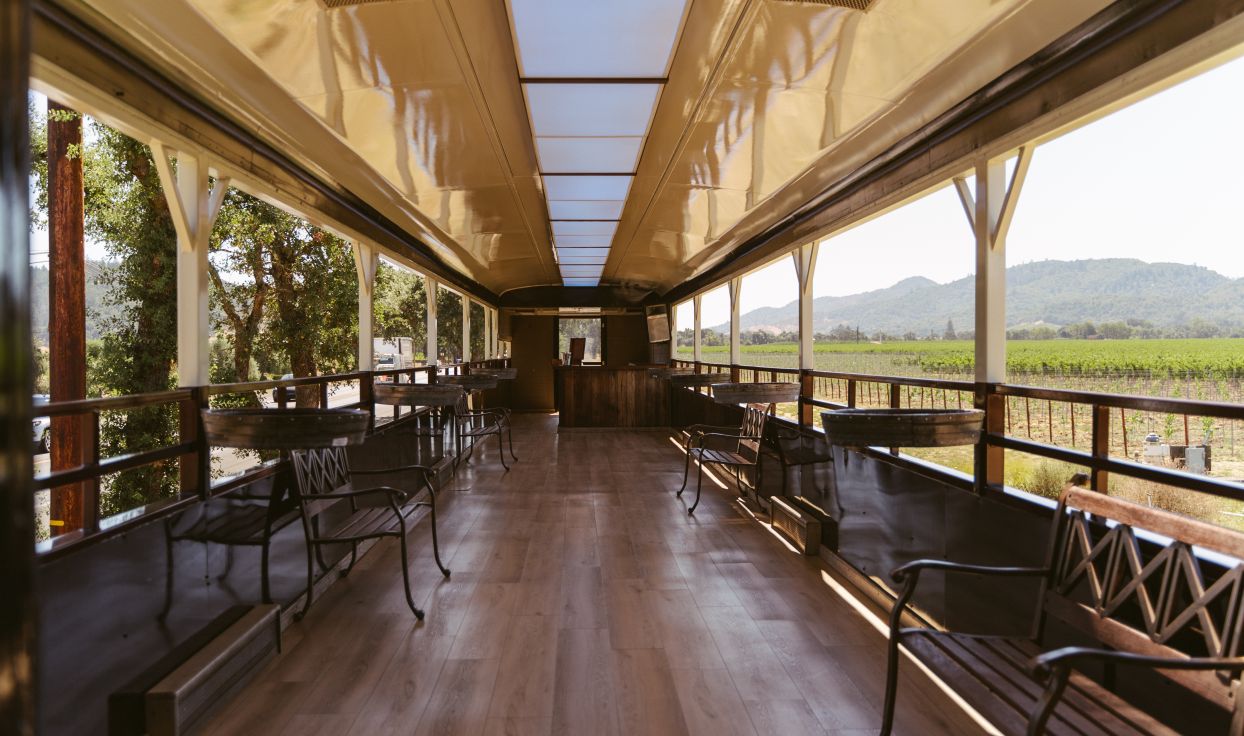 A photo from within a cabin of the Napa Valley Wine Train. Where there are normally windows, the space appears to be wide open. Wooden chairs and benches are lined against the sides of the cabin.