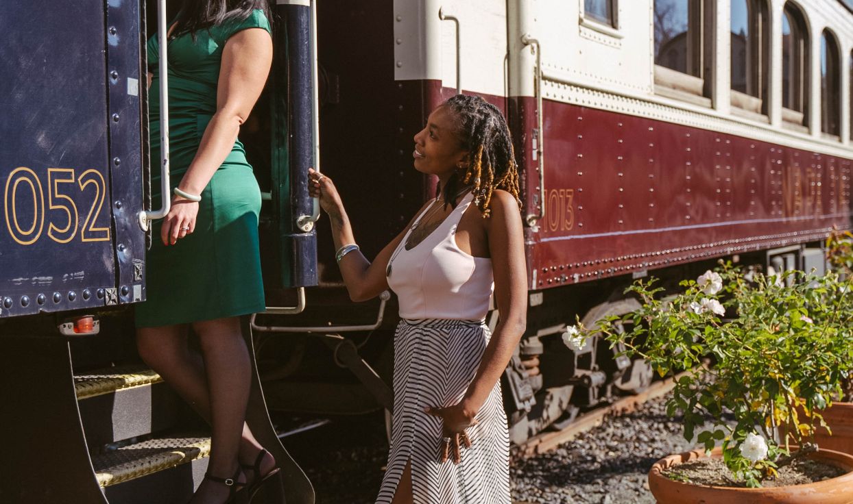 Two women look at each other, and are posed boarding the Napa Valley Wine Train. The woman on the left stands on the stairs to the train's cabin, wearing a green dress. The woman on the right is standing on the ground, weaking a flowy white pattered dress.