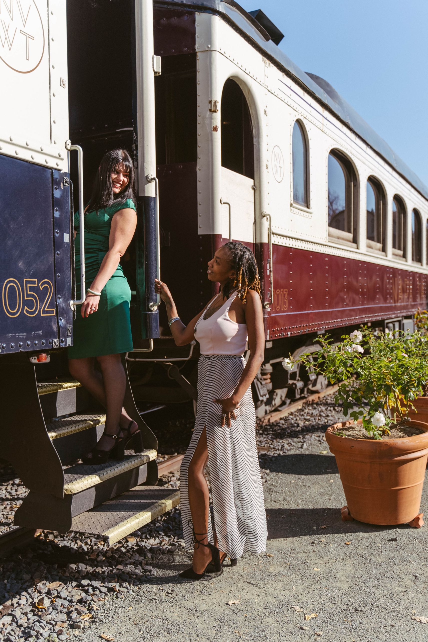 Two women look at each other, and are posed boarding the Napa Valley Wine Train. The woman on the left stands on the stairs to the train's cabin, wearing a green dress. The woman on the right is standing on the ground, weaking a flowy white pattered dress.