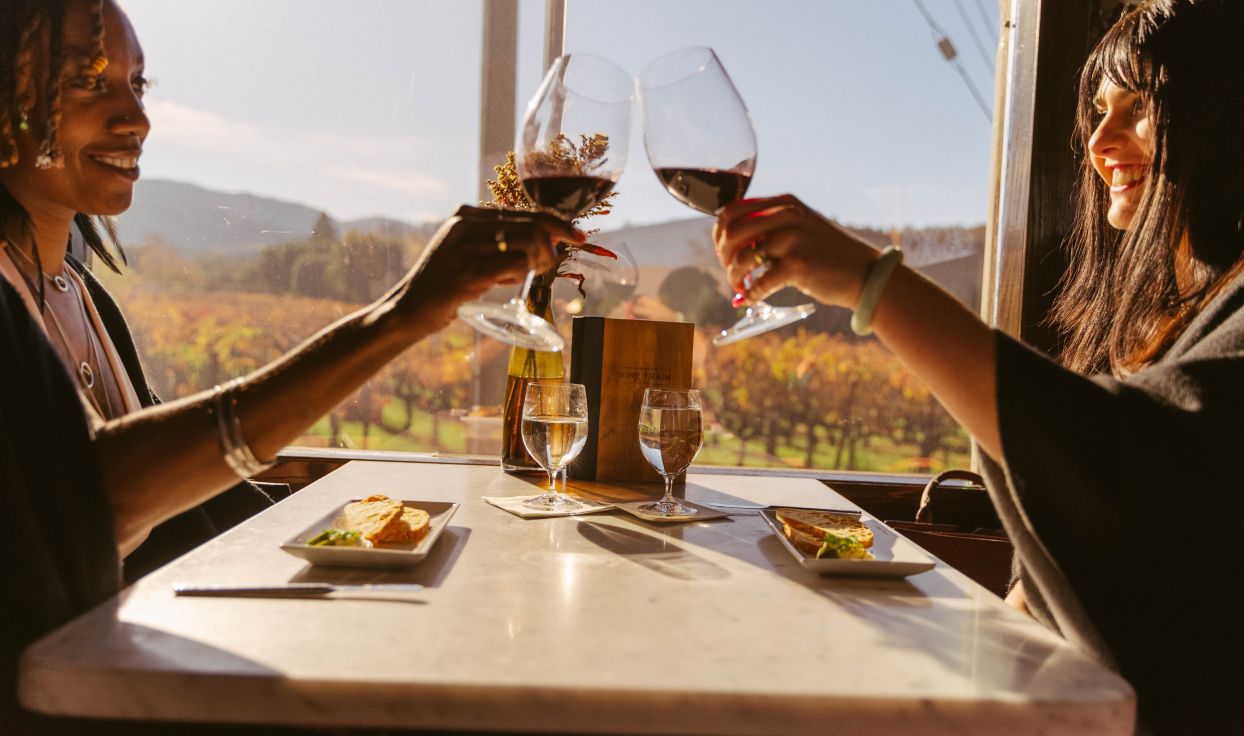 Two people sit at a table onboard the Napa Valley Wine Train. They each hold a glass of red wine, clicking their glasses together over their table.
