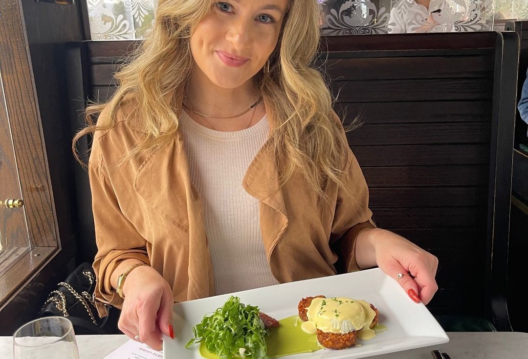 A blonde woman poses for the camera, displaying a plate of food in her hands. The plated food consists of leafy greens and an egg with a yellow sauce.