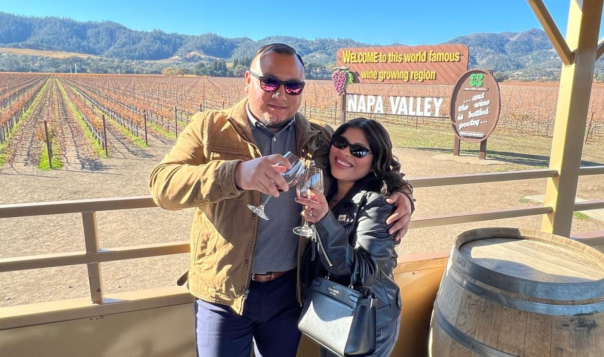 A man and woman wearing sunglasses clink their glasses of red wine together. They are posed in front of the Napa Valley wooden sign and vineyard.