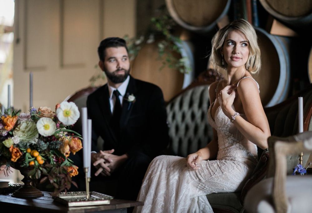A man and woman sit next to each other on a tufted couch. The man wears a black suit and tie, with a white rose boutonniere on his label. The woman wears a long fitted and beaded wedding gown. They are both gazing towards the right. A bouquet of orange and white flowers sits on table to the left, surrounded by tall white candles in golden candlesticks.
