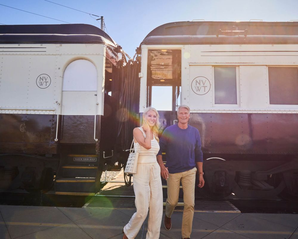 A man and woman are pictured smiling and holding hands outside of the Napa Valley Wine Train cabins. The woman is wearing an all off-white coloured outfit. The man wears a blue longsleeve and khakis.