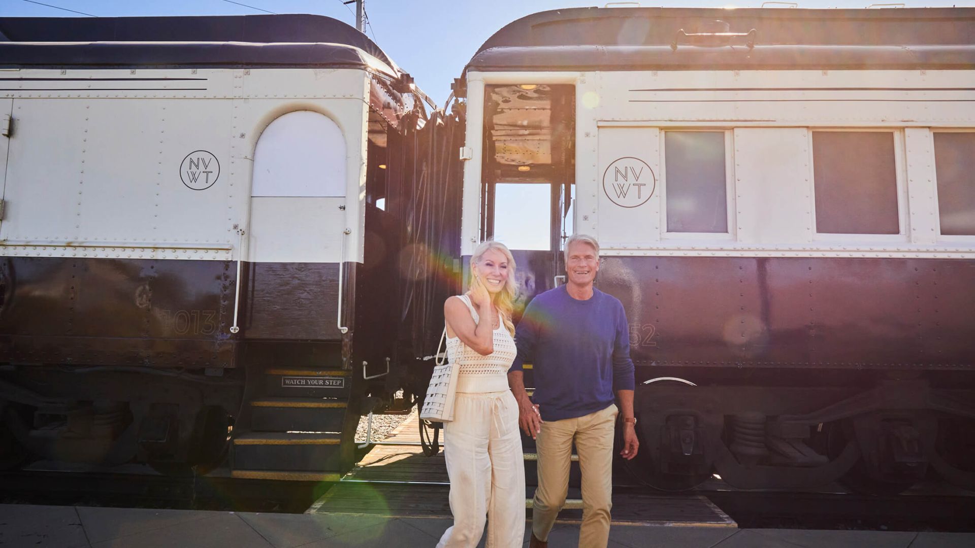 A man and woman are pictured smiling and holding hands outside of the Napa Valley Wine Train cabins. The woman is wearing an all off-white coloured outfit. The man wears a blue longsleeve and khakis.