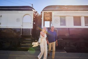 A man and woman are pictured smiling and holding hands outside of the Napa Valley Wine Train cabins. The woman is wearing an all off-white coloured outfit. The man wears a blue longsleeve and khakis.