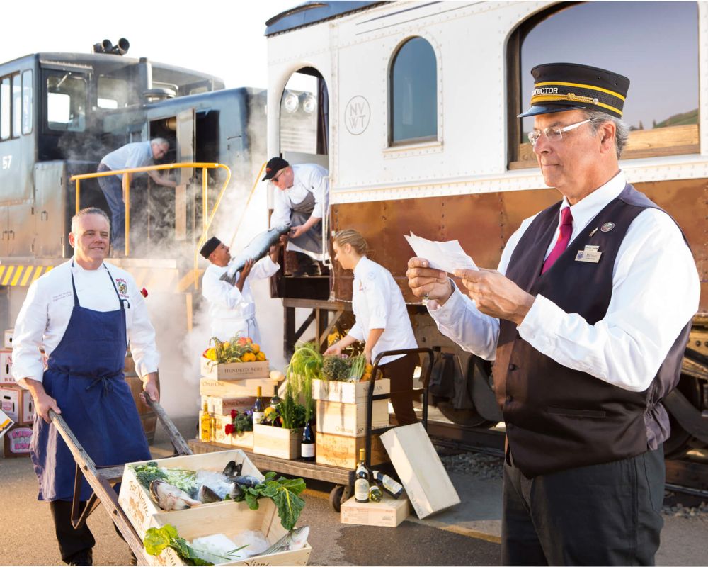 A scene of Napa Valley Wine Train staff posed outside of the Napa Valley Wine Train. To the left, a man wearing a blue apron wheels fresh produce and fish in a wooden wheelbarrow. To the right, the train's conductor reads from a paper in his hand. Behind the two men are four staff members, three which are loading fish and produce onto the train, and an engineer looking into the train's engine doors.