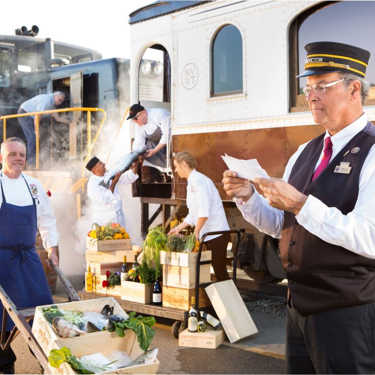 A scene of Napa Valley Wine Train staff posed outside of the Napa Valley Wine Train. To the left, a man wearing a blue apron wheels fresh produce and fish in a wooden wheelbarrow. To the right, the train's conductor reads from a paper in his hand. Behind the two men are four staff members, three which are loading fish and produce onto the train, and an engineer looking into the train's engine doors.