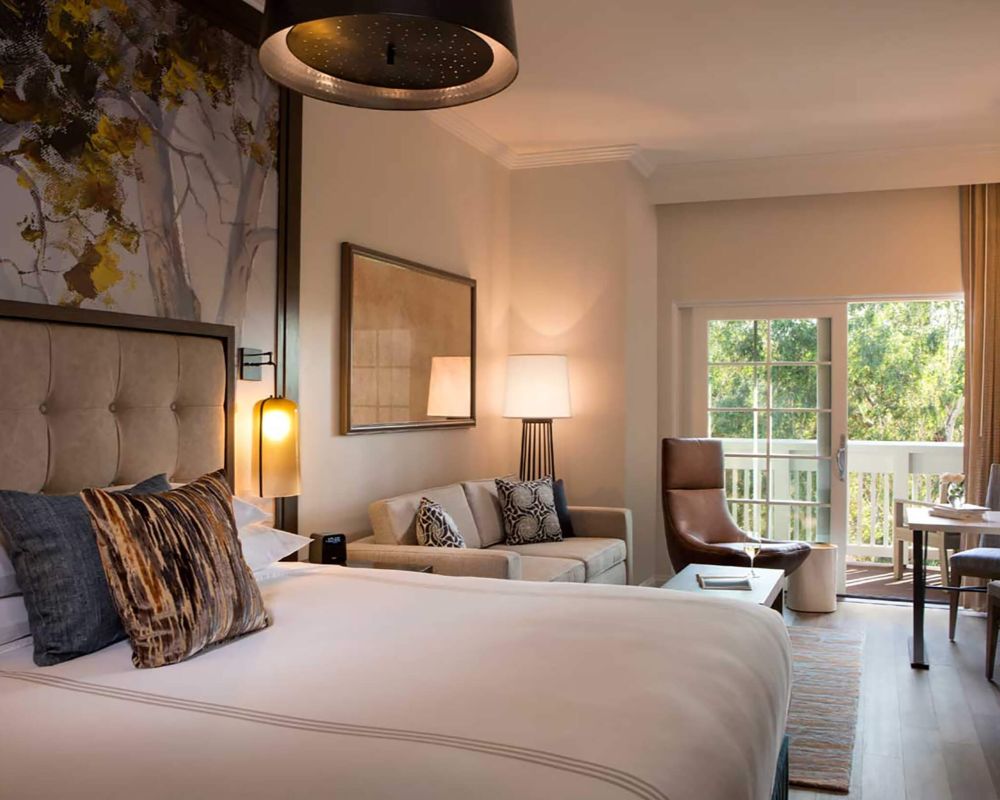 A photo of a hotel room. There is a large bed dressed in white linens and dark, patterned pillows, and a cushioned headboard. Towards the centre, a beige couch with more dark patterned pillows. To the right, more furniture, such as a coffee table, a leather lounge chair, two dining chairs, and a dining table.