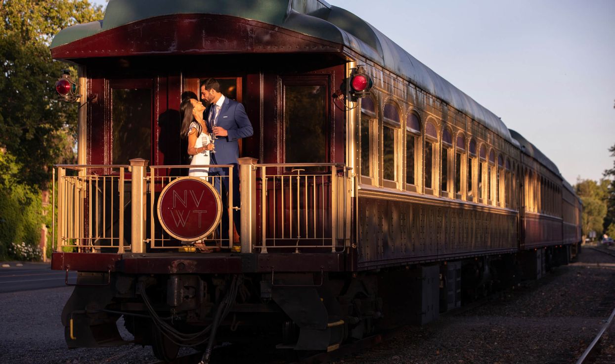 The outside of the Napa Valley Wine Train. A man in a blue suit and a woman in a white dress stand on the back of the train's caboose, both holding champagne flutes.