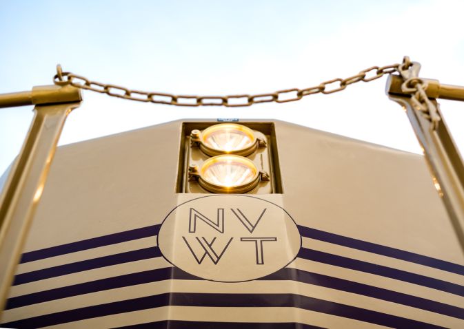 The front of the Napa Valley Wine Train, taken from a low perspective. Two headlights are illuminated, with the Napa Valley Wine Train logo sitting underneath them. There is a golden chain link positioned in front of the train.